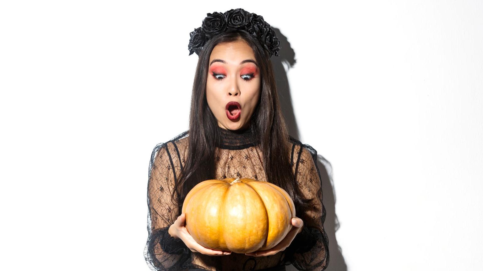 Image of amazed asian girl looking at big pumpkin fascinated, wearing gothic dress and black wreath, standing over white background